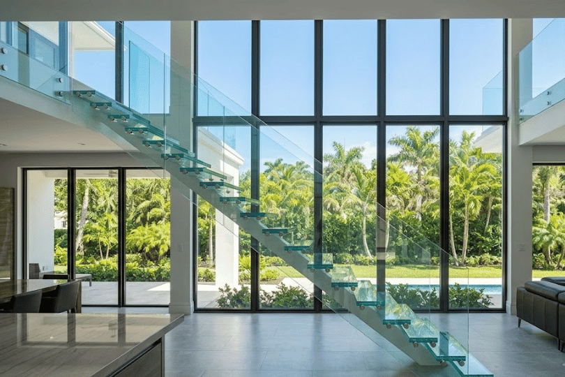 Modern interior home design featuring a floating glass staircase next to a floor-to-ceiling glass wall overlooking a tropical garden.